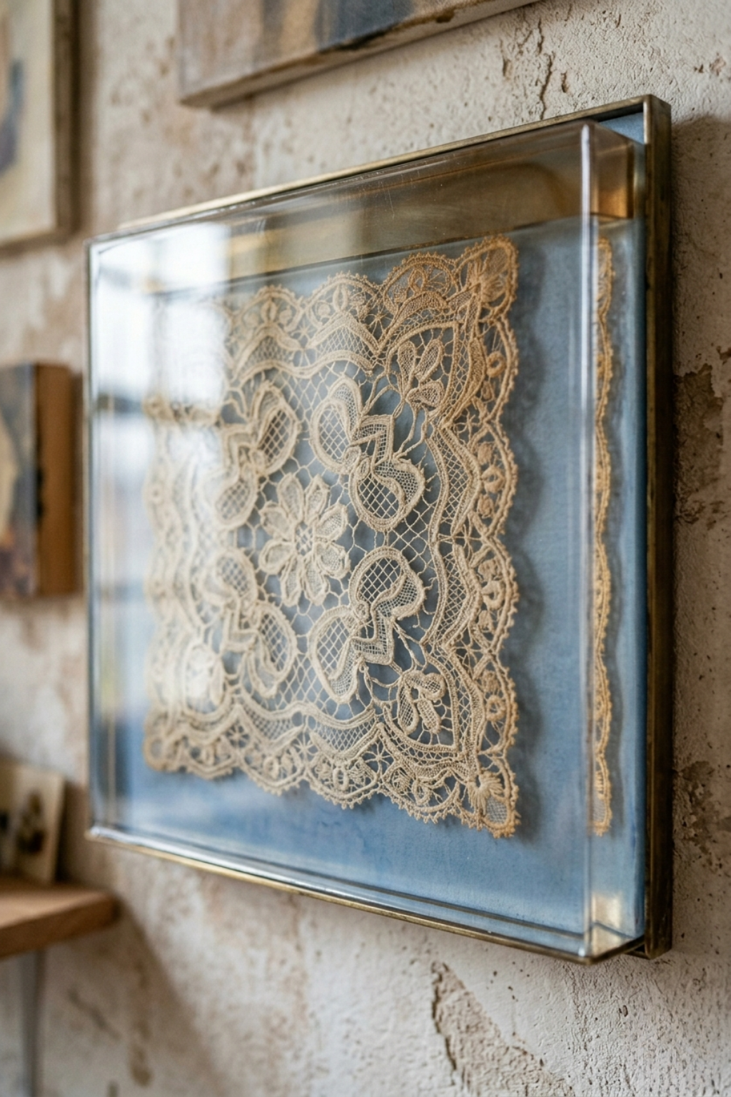 Vintage handmade lace doily displayed in a gold shadow box frame on a rustic textured wall.