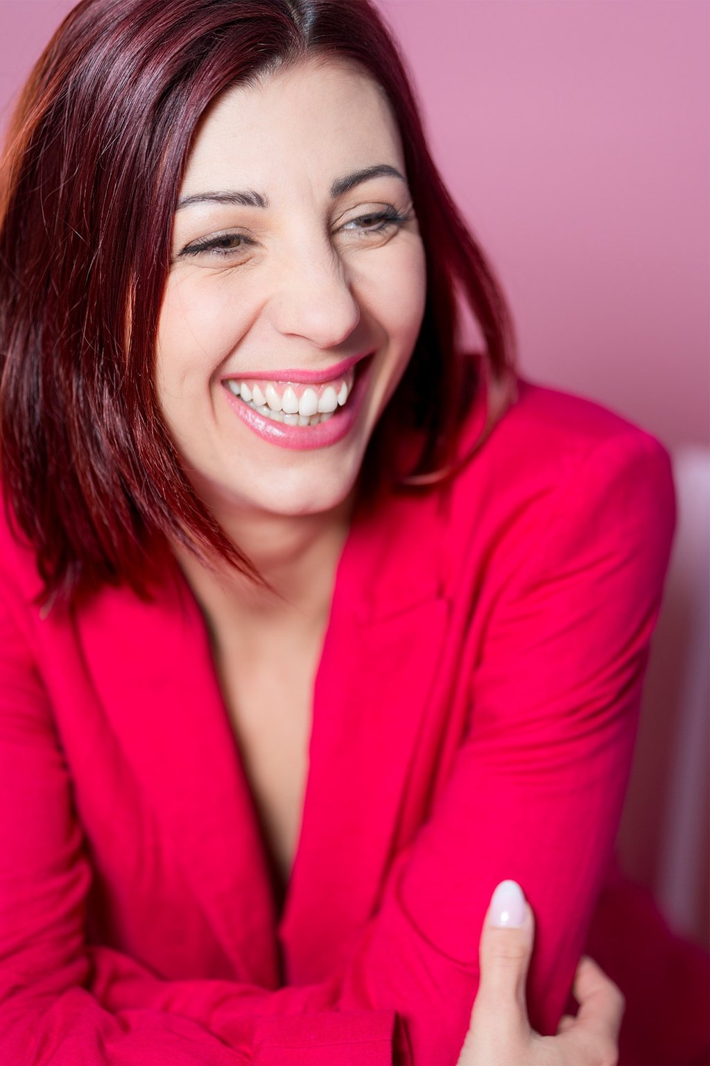Portrait d’une comédienne qui rit, en veste rose sur fond rose, sourire naturel et expressif.
