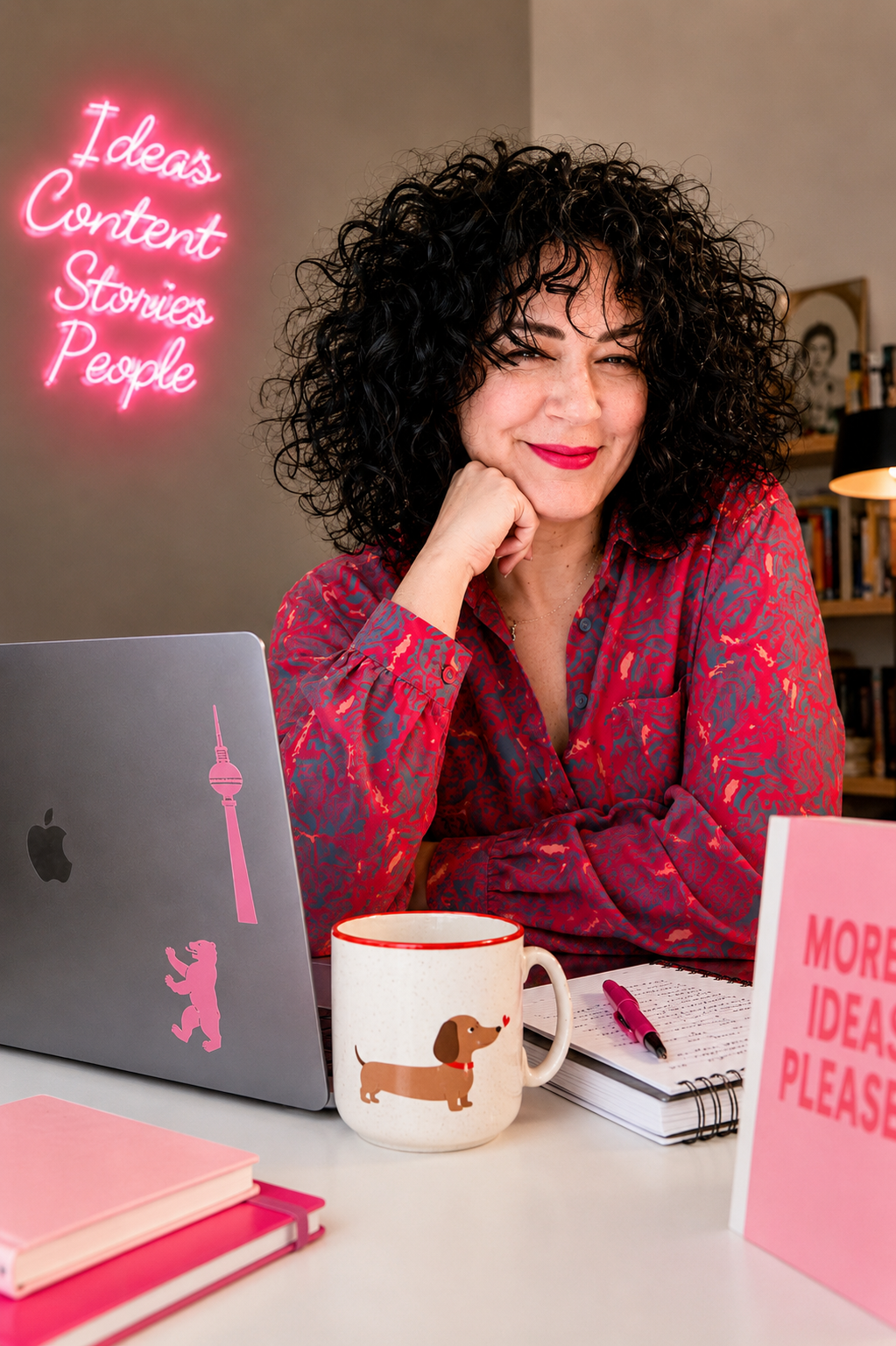 Content creator with curly hair working at a desk with a laptop and neon sign.