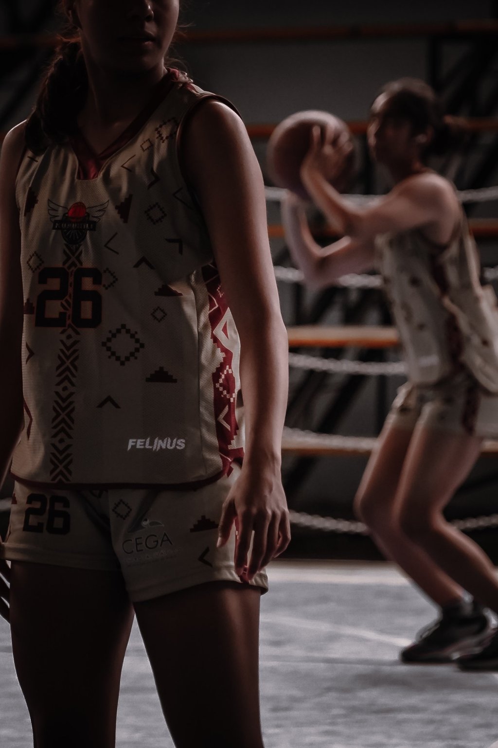 A female basketball player in a custom patterned jersey stands on court while a teammate shoots a ball.