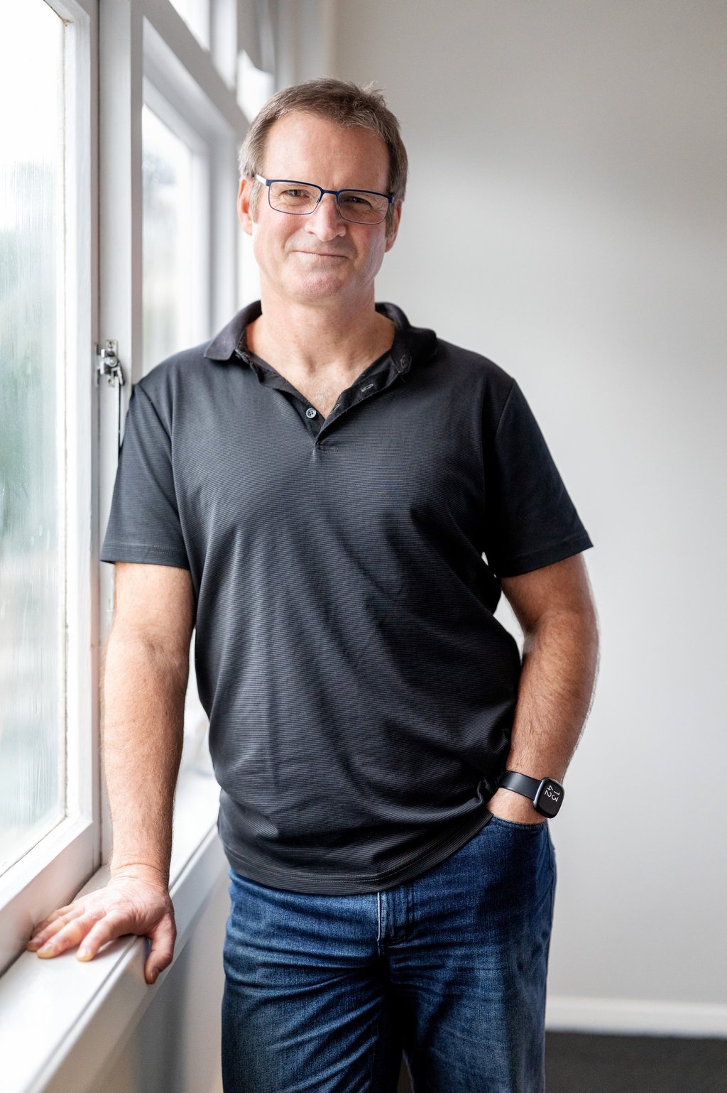 Photo of a man wearing a glasses, a black polo shirt and jeans, leading on a window sill and smiling