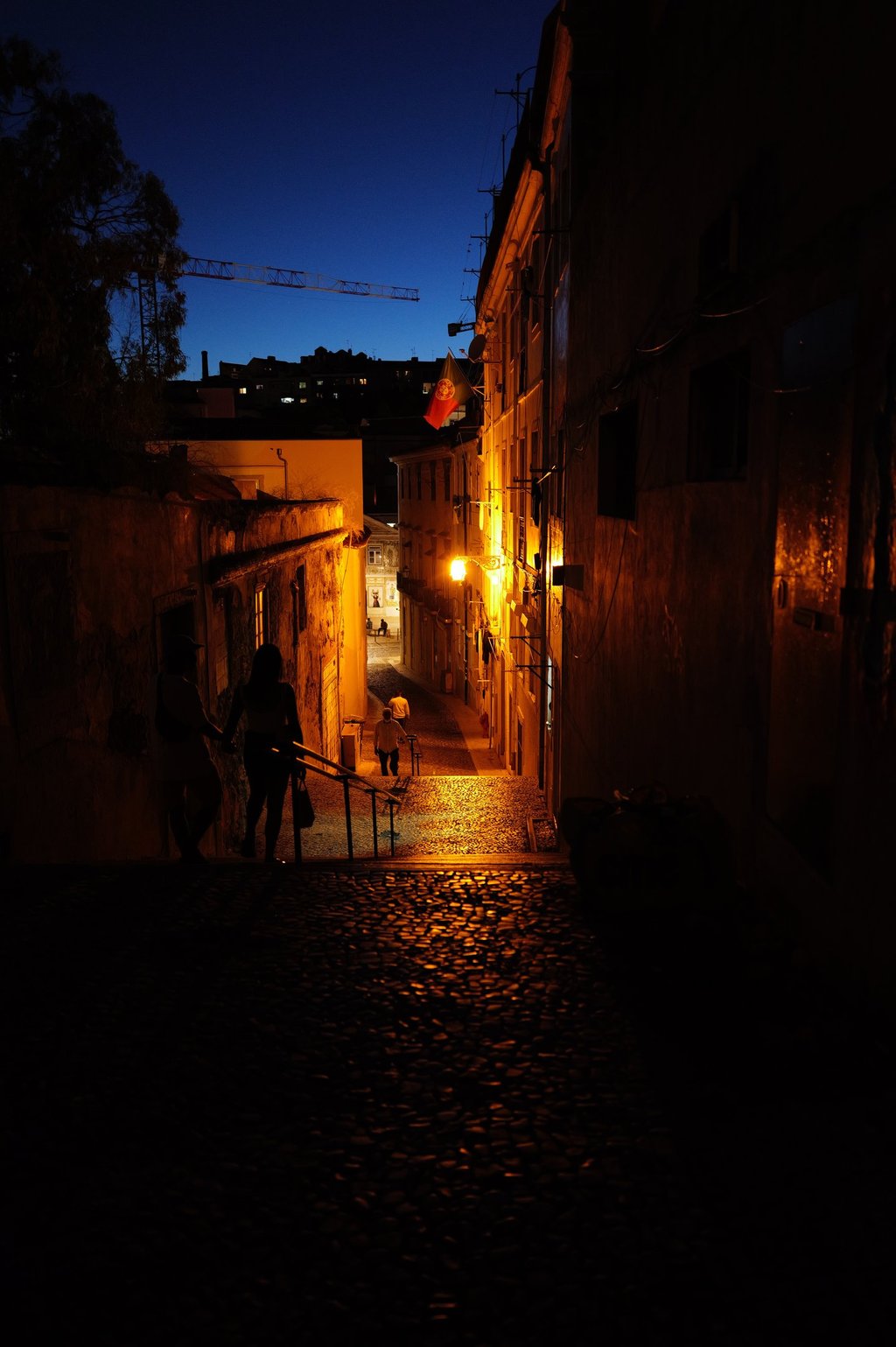 Night cobblestone alley lit of Lisbon by warm streetlights with walking silhouettes, By ACAT Photos