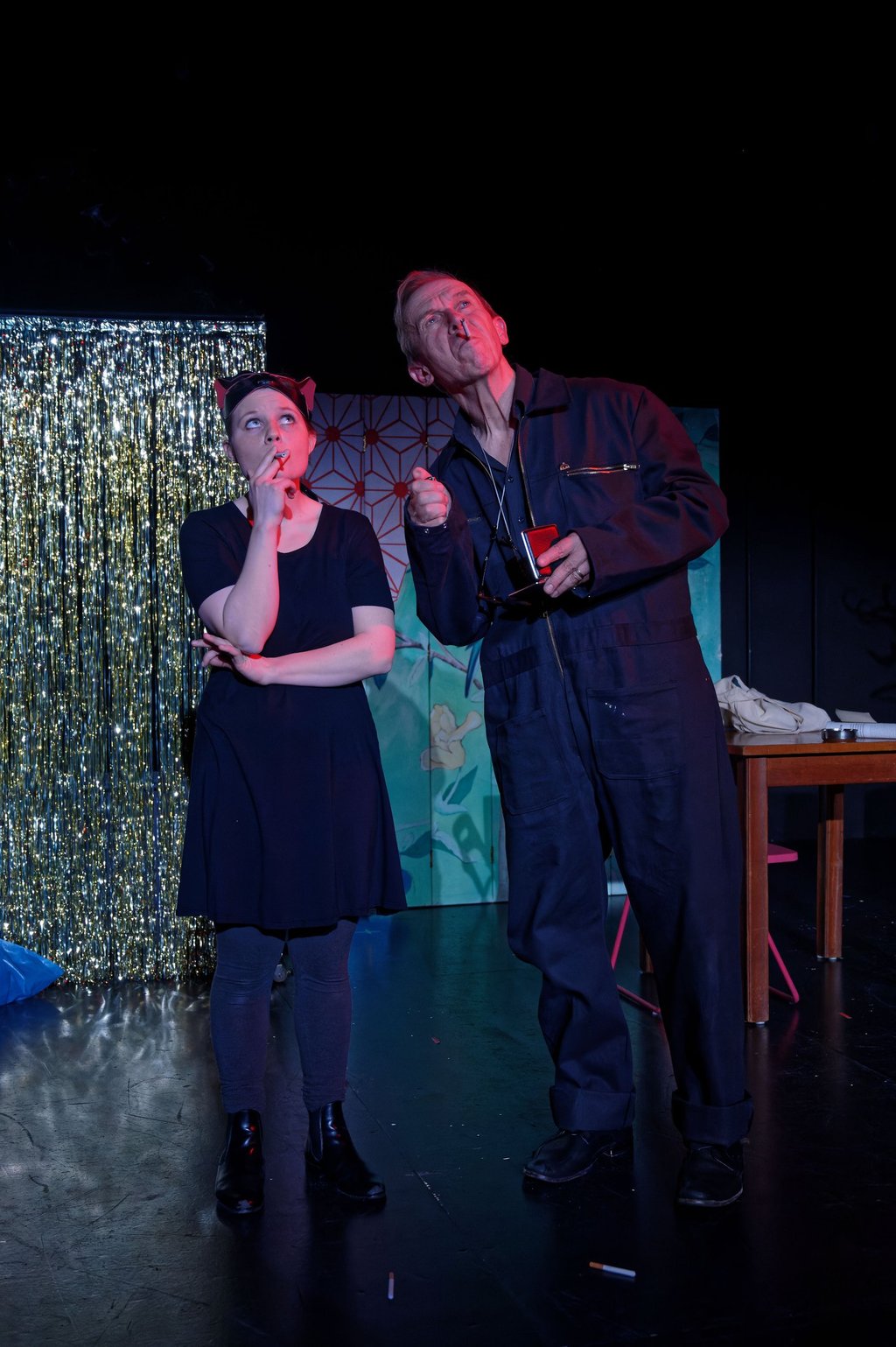 Zwei Schauspieler:innen rauchen vor einem glitzernden Vorhang.
