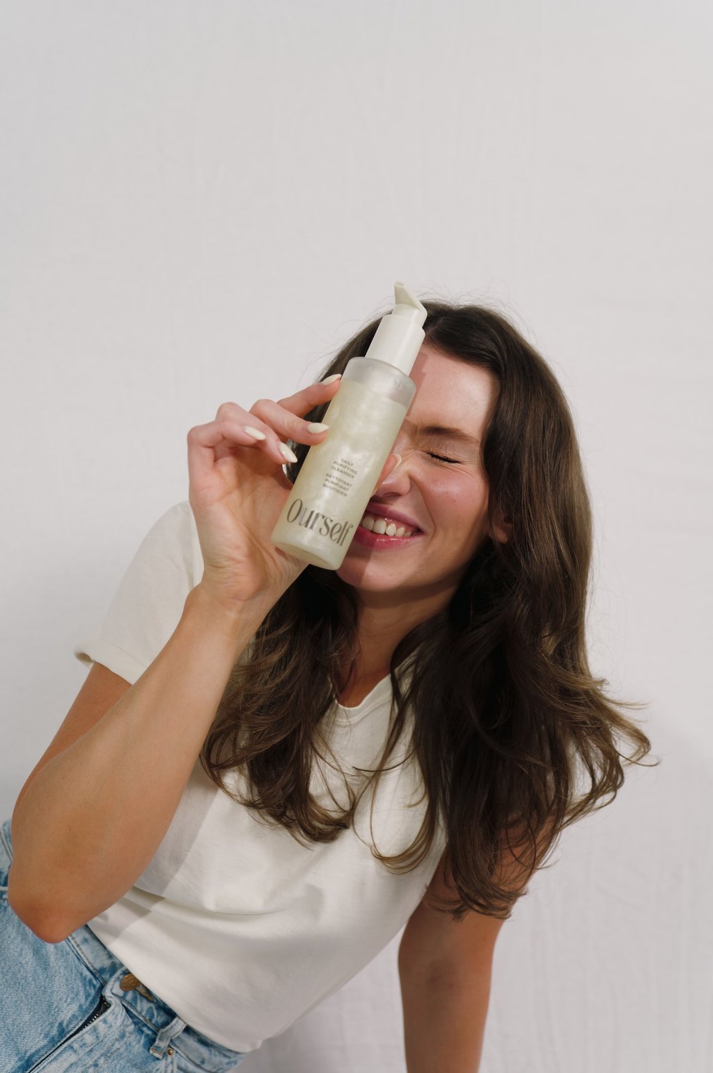 A smiling woman holding a bottle of Ourself daily purifying facial cleanser against her face.