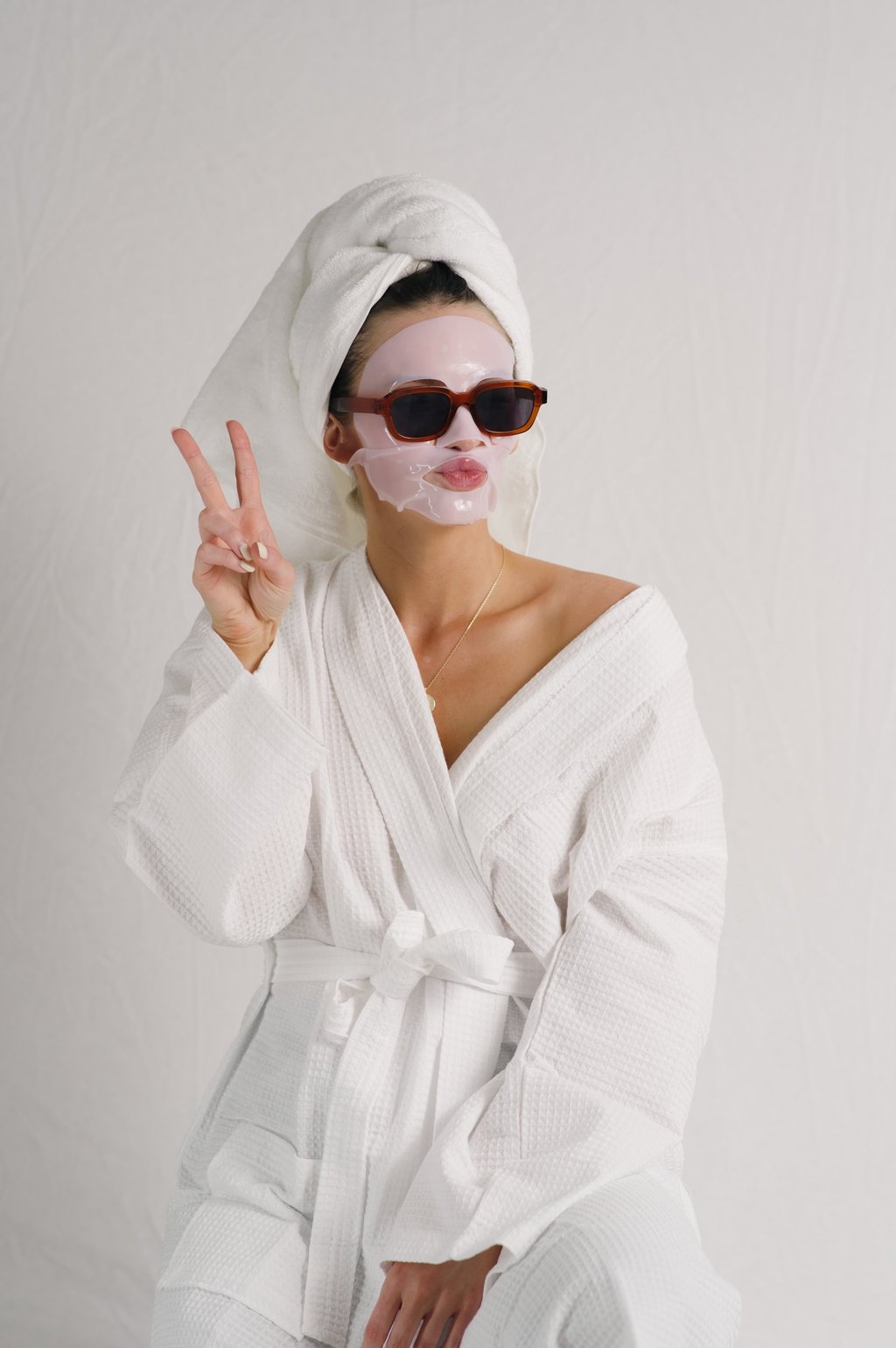 Woman in a white bathrobe and towel wrap wearing a pink sheet face mask and sunglasses while making a peace sign.