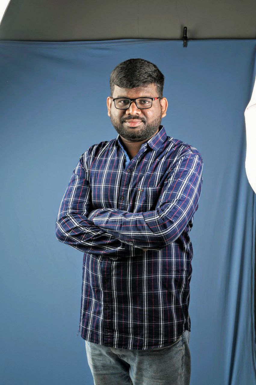 Professional portrait of a man in a plaid shirt with arms crossed against a blue backdrop.