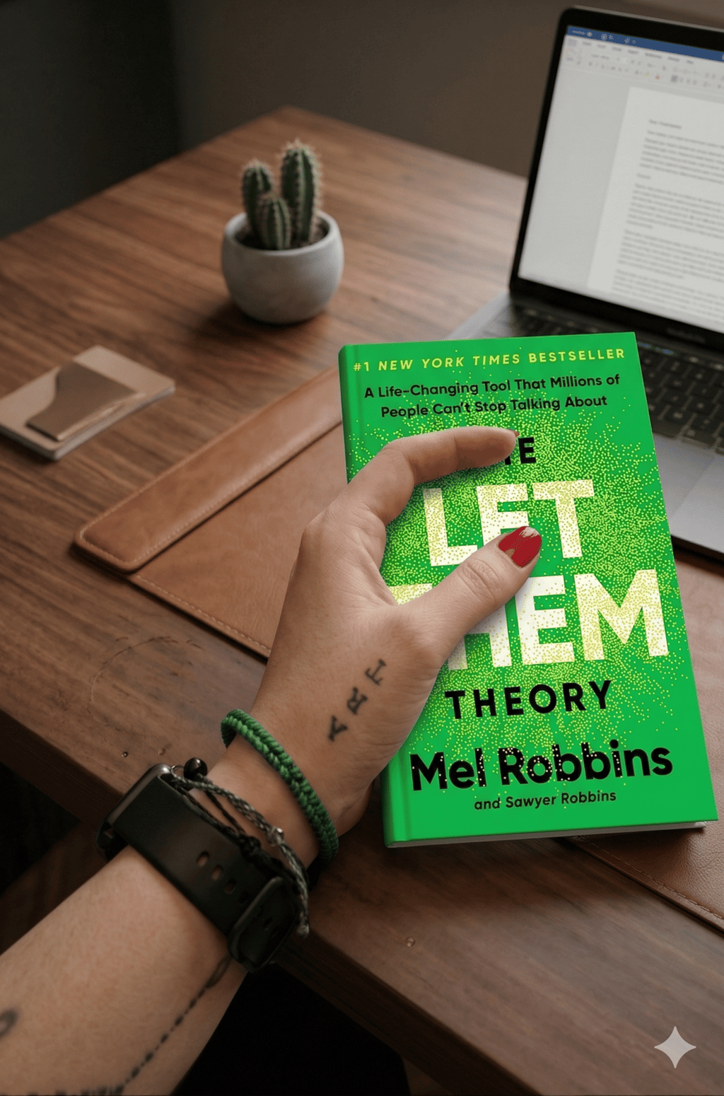 A person holding The Let Them Theory book by Mel Robbins at a wooden desk with a laptop.