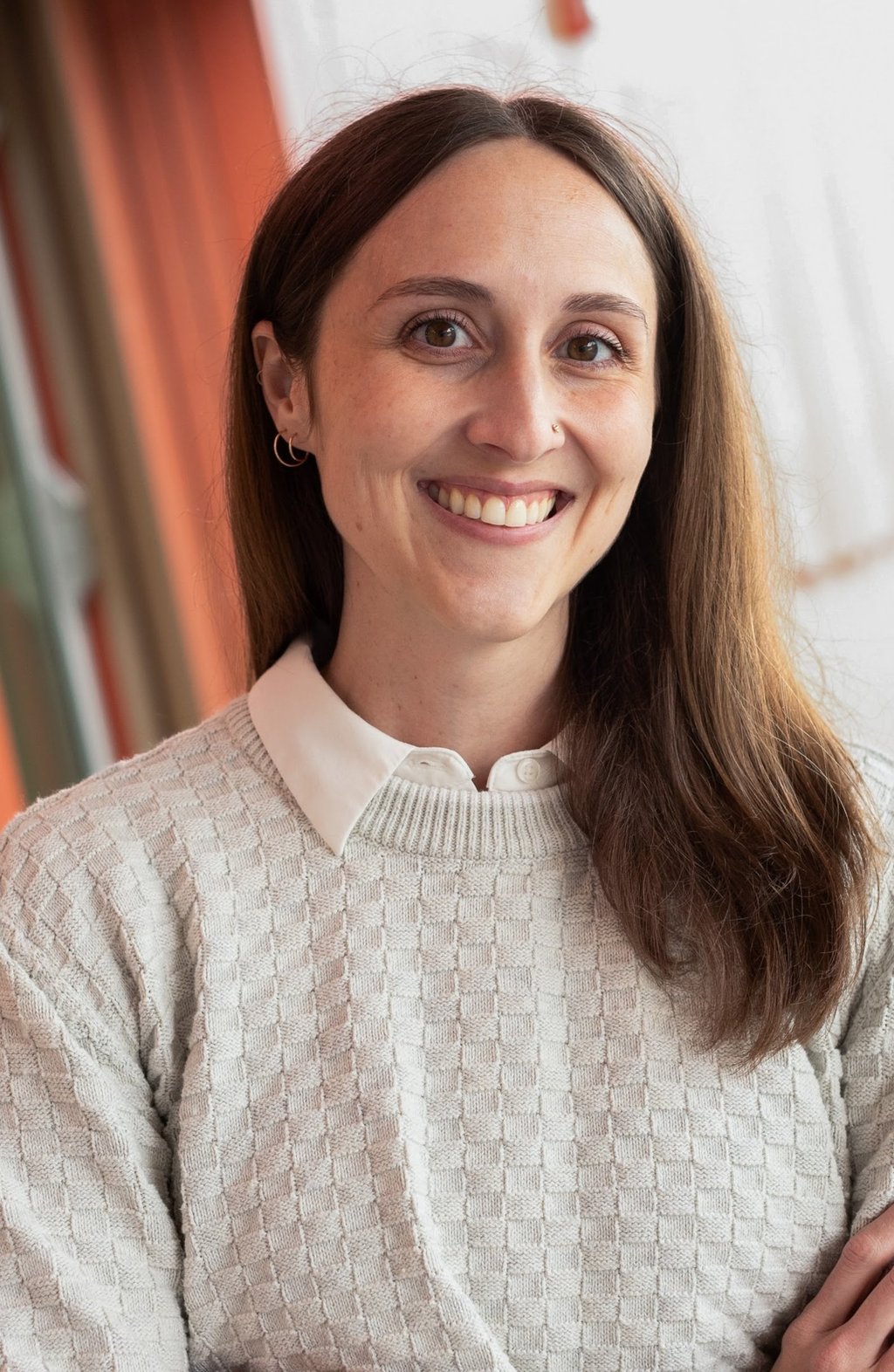 A smiling woman with long brown hair smiling, wearing a cream sweater over a white collared shirt 