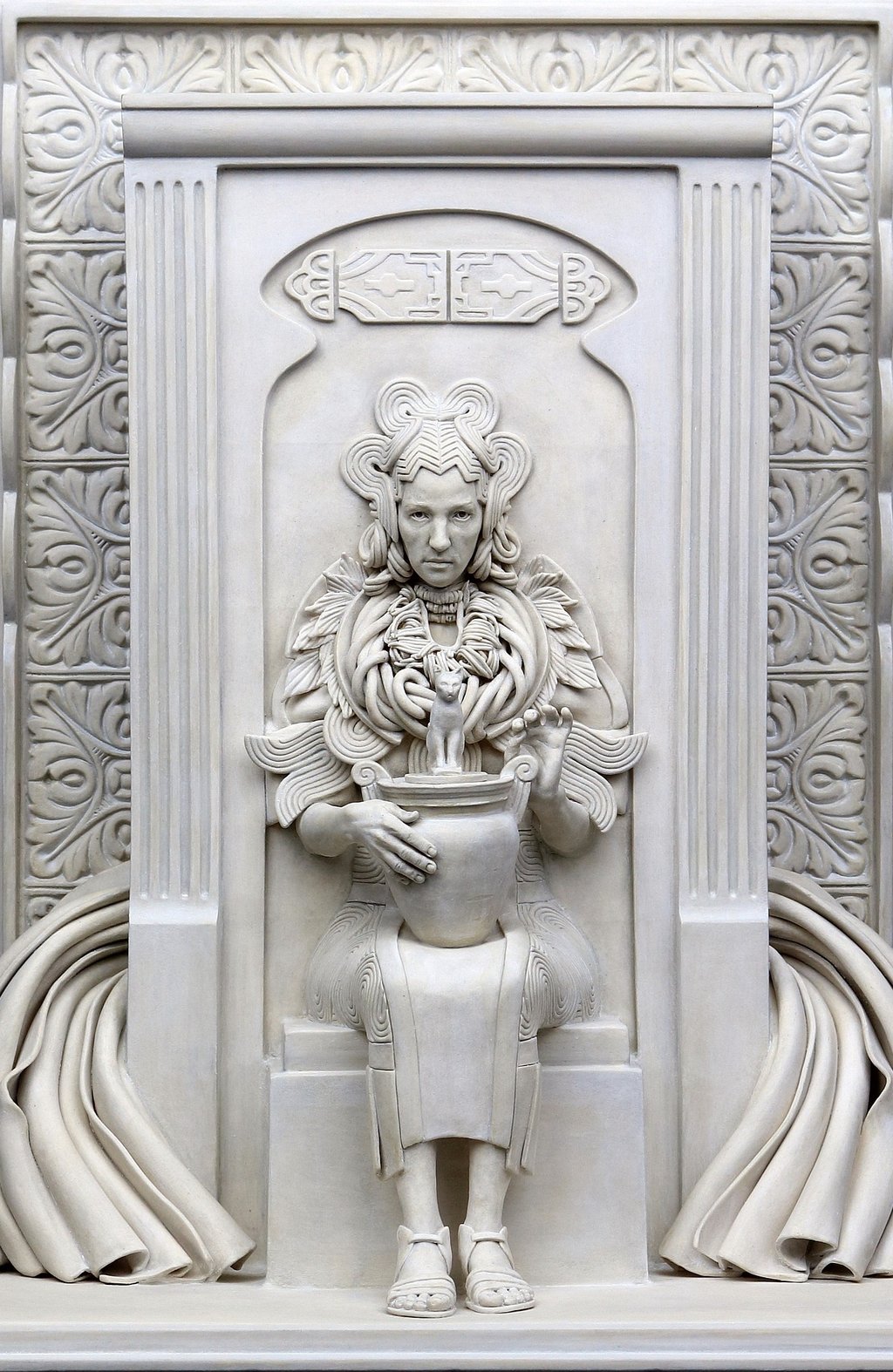 Ceramic sculpture of woman embedded in wall, wearing heavy jewelry and elaborate dress, holding Egyptian cat urn; closeup.