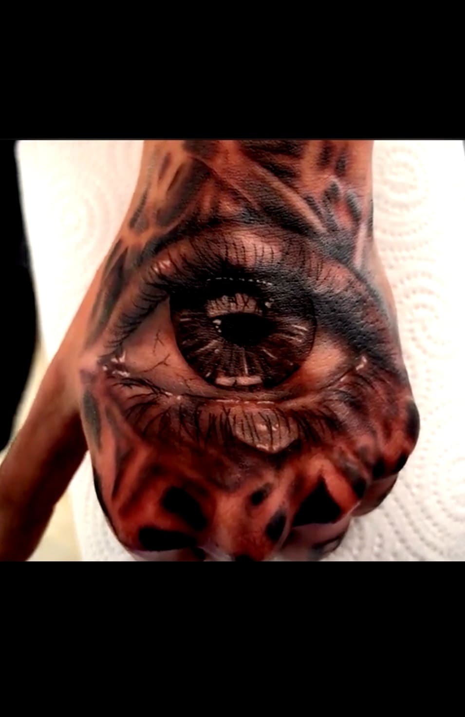 Realistic black and grey eye tattoo on a hand with intricate pupil details and surrounding dark patterns.