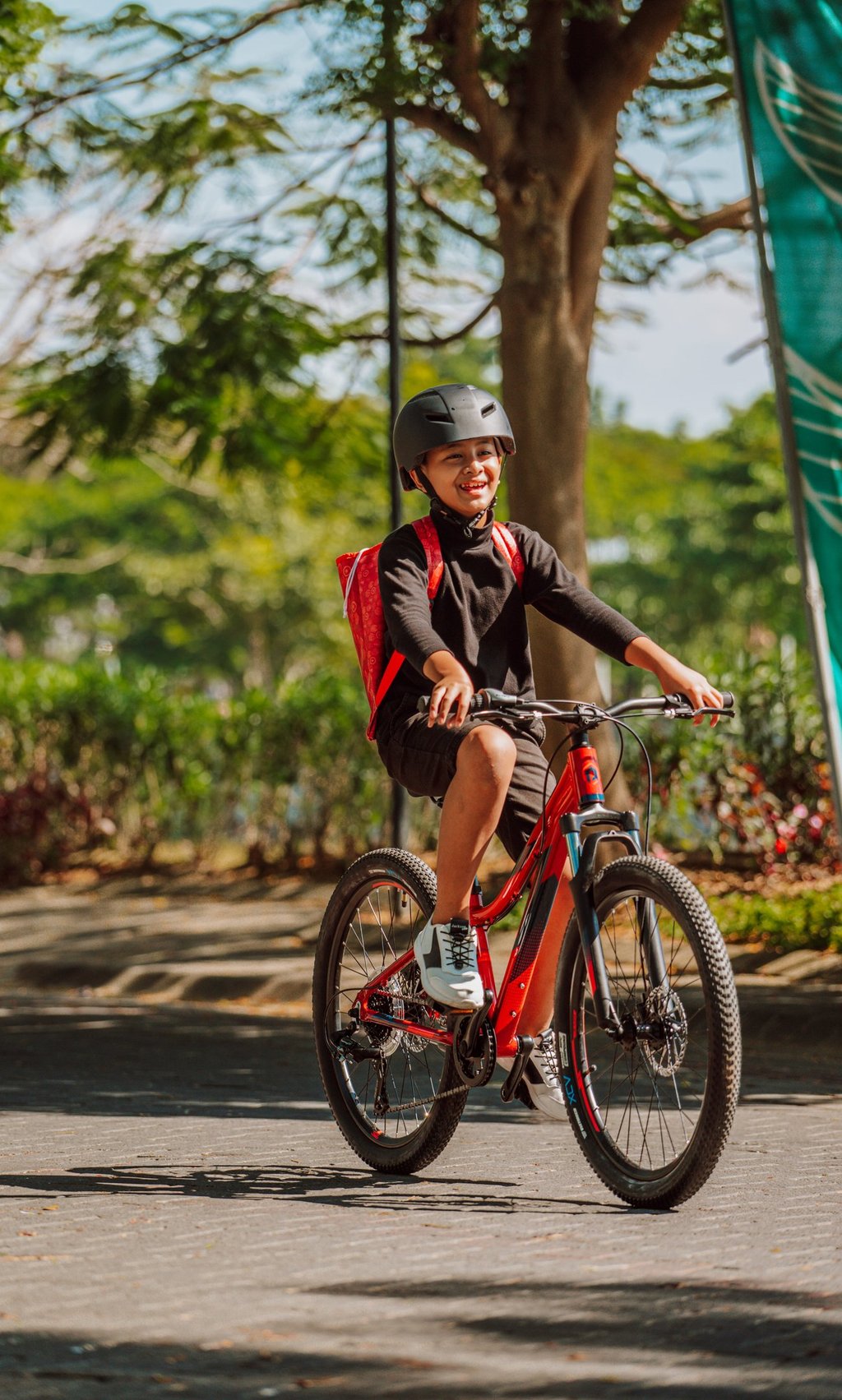Smiling young boy wearing a helmet and backpack riding a red mountain bike on a sunny park path.