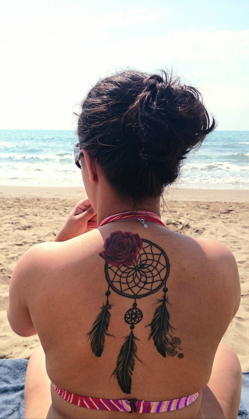 Mulher de costas na praia, com tatuagem de rosa e filtro dos sonhos, símbolo de memória ancestral