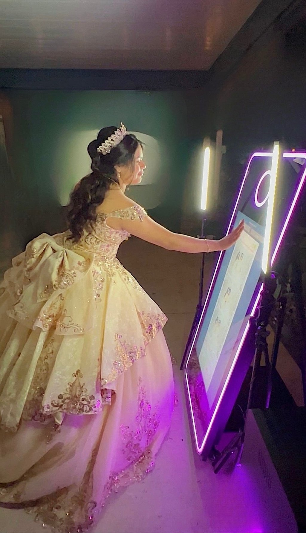 Elegant quinceañera interacting with illuminated mirror booth in a sparkling blush and gold gown.