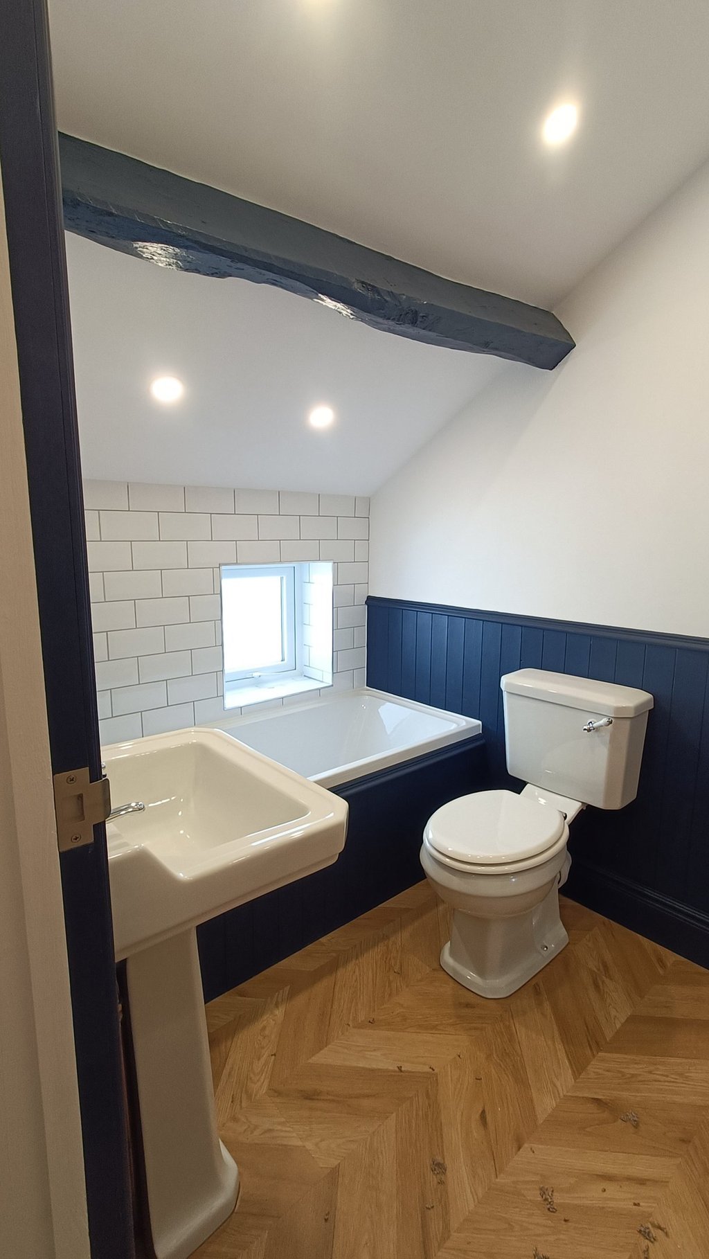 Modern attic bathroom with white bathtub, pedestal sink, and blue wood paneling.