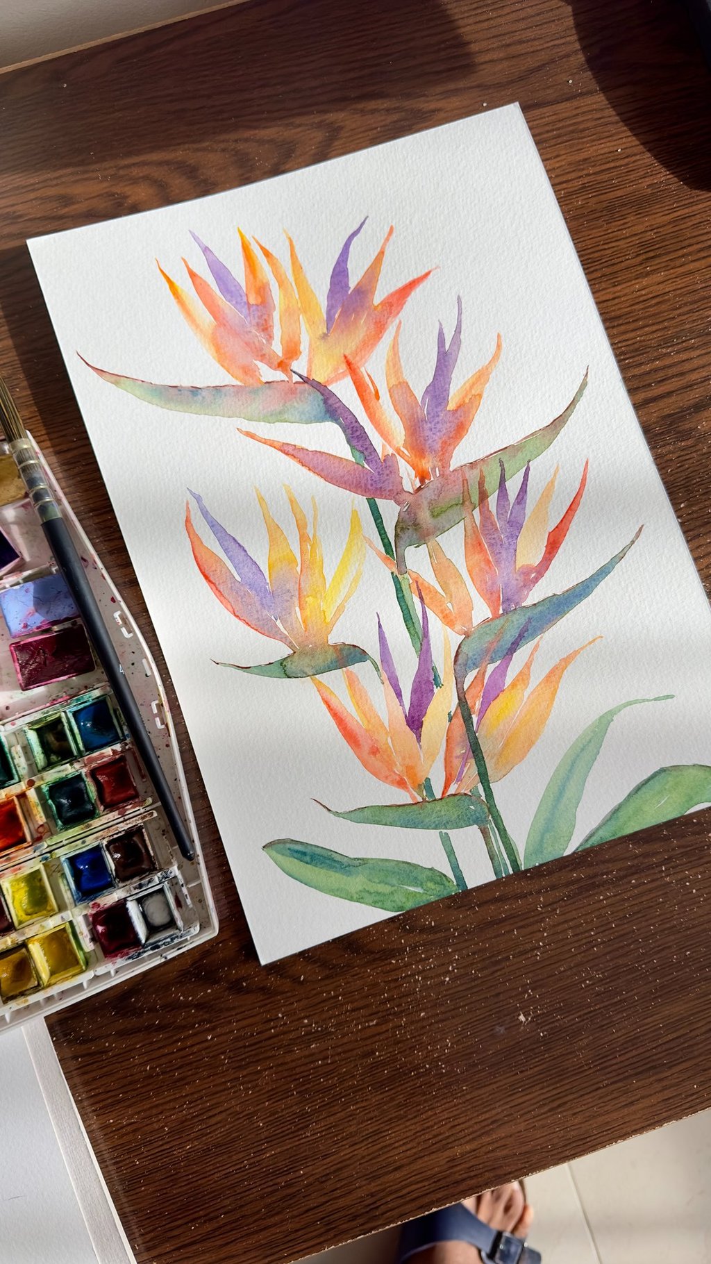 Vibrant watercolor painting of a Strelitzia flower with orange and blue petals resting on an artist's workspace.
