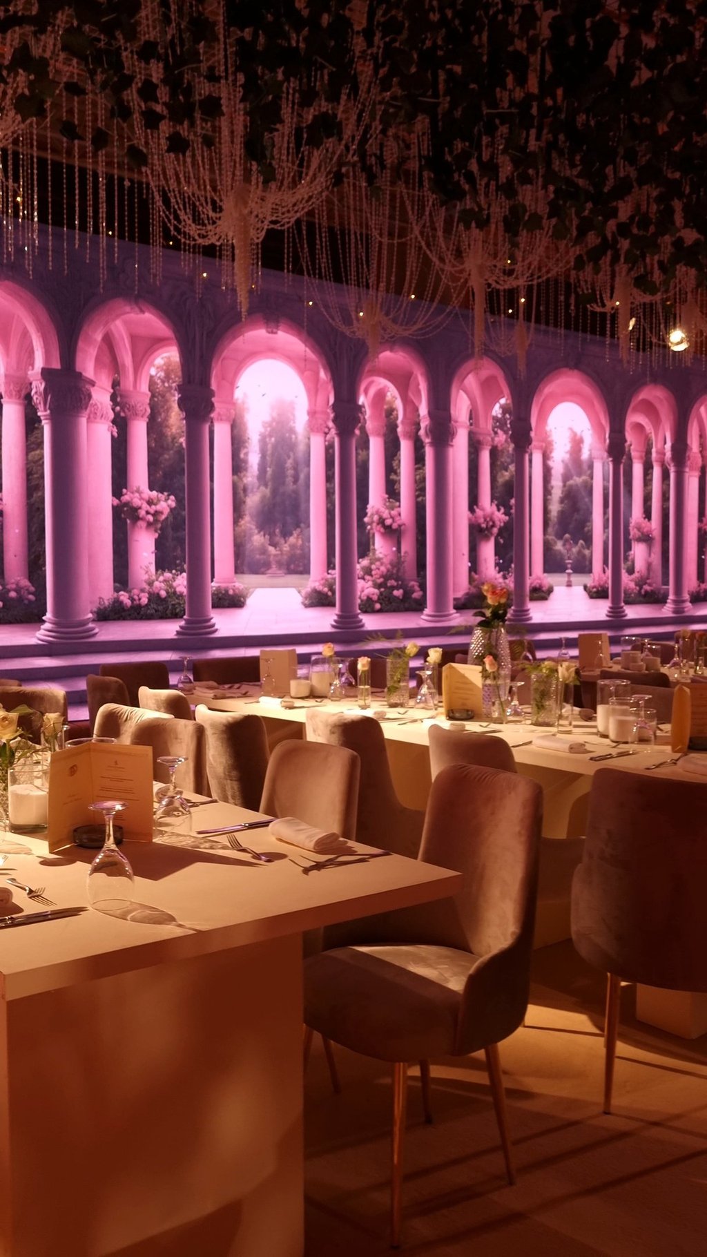 a wedding with a long table and chairs and large led screen in the back with roman arches and garden