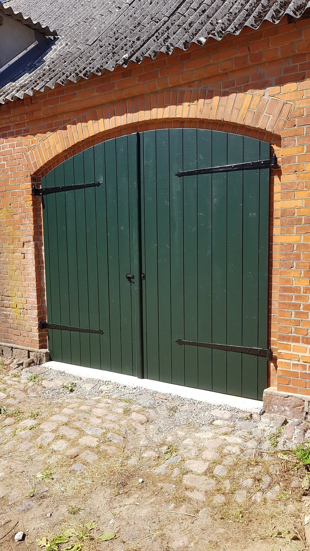 Mørkegrønne buede træporte med sorte metalhængsler, indsat i en rustik rød murstensvæg.