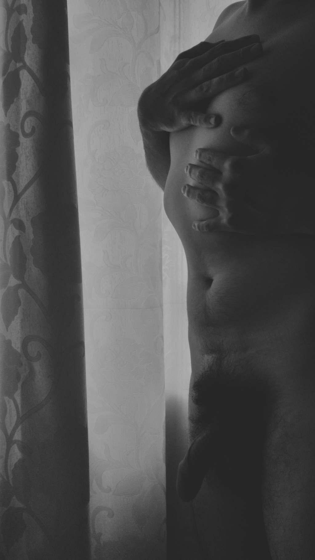 A black-and-white artistic male nude self-portrait in front of a curtain.
