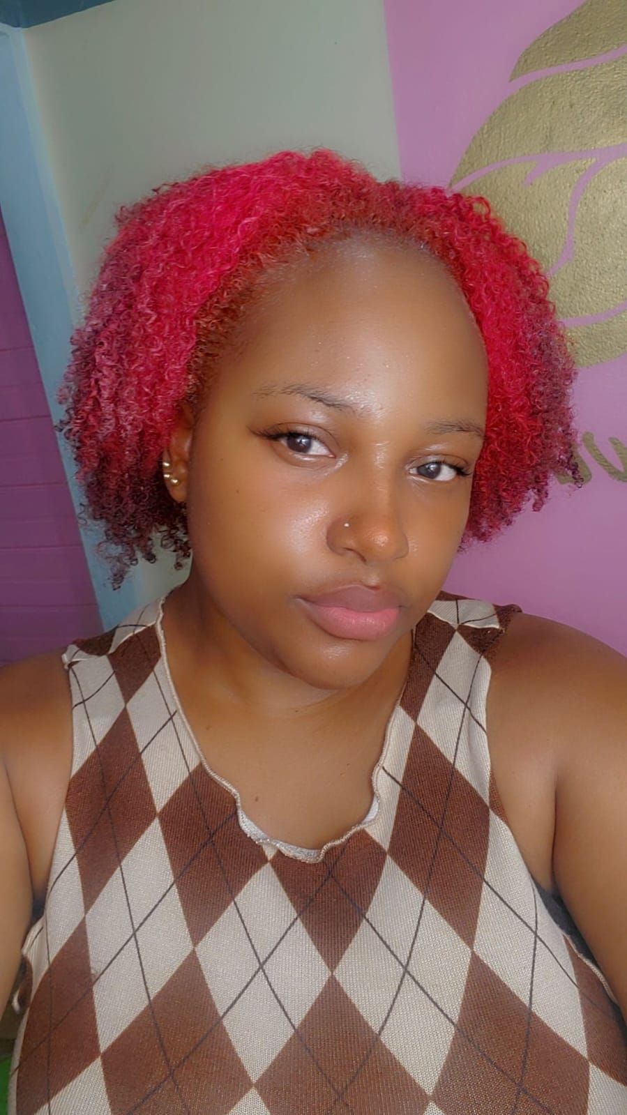 A young woman with rosy pink natural curls, styled in a short, voluminous shape. Her glowing skin, subtle makeup, and nose pi