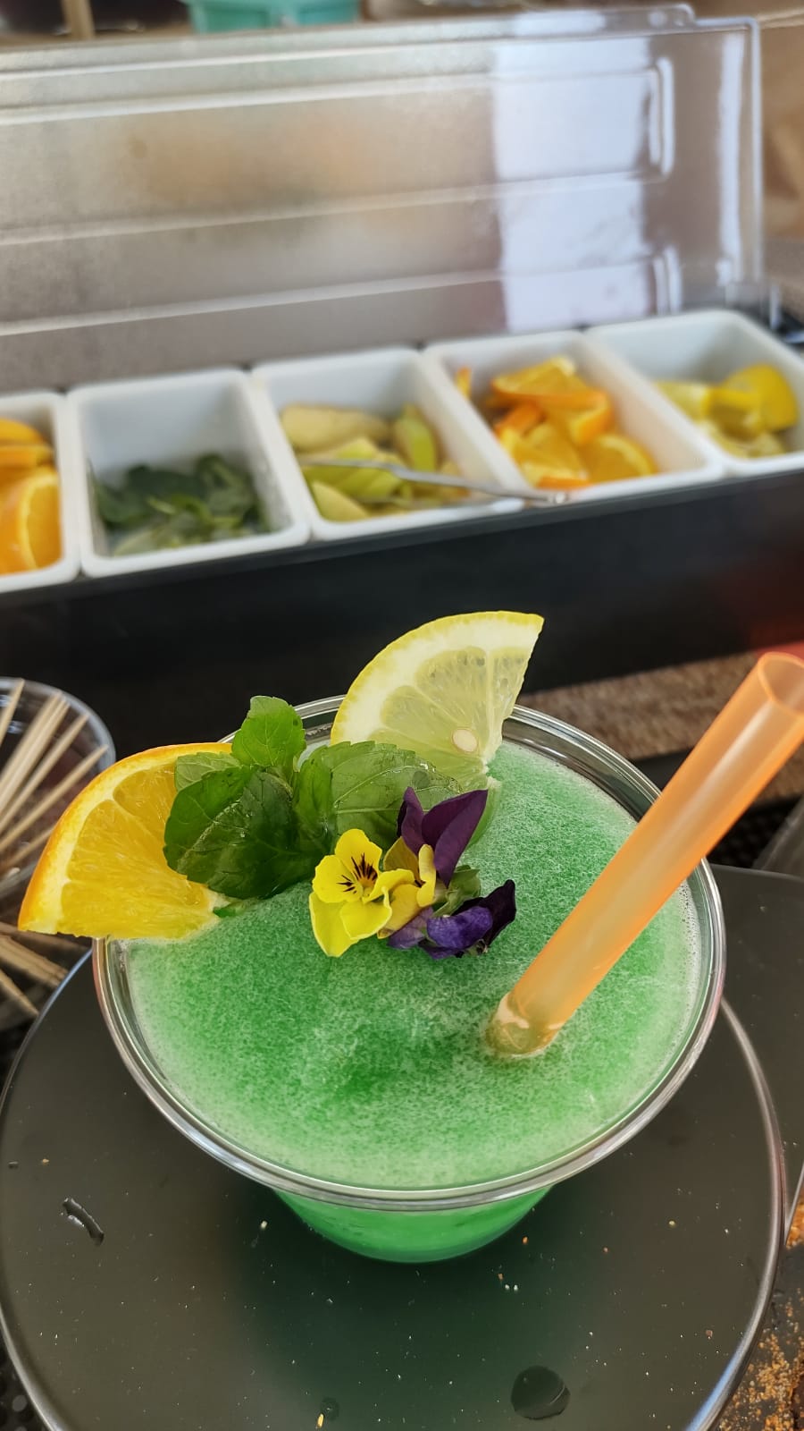 Coctel frappé verde adornado con menta fresca, rodajas de cítricos y flores comestibles