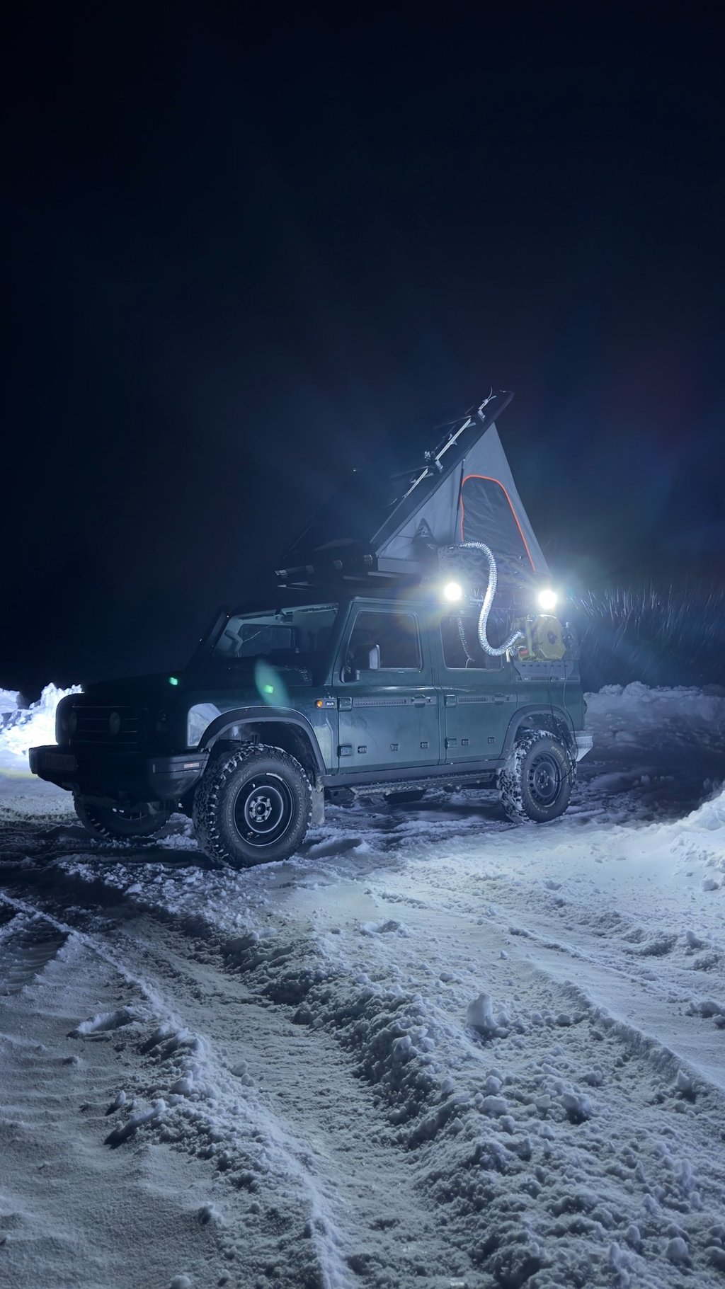 Off-road 4x4 vehicle with a rooftop tent and diesel heater setup for winter camping in the snow at night.