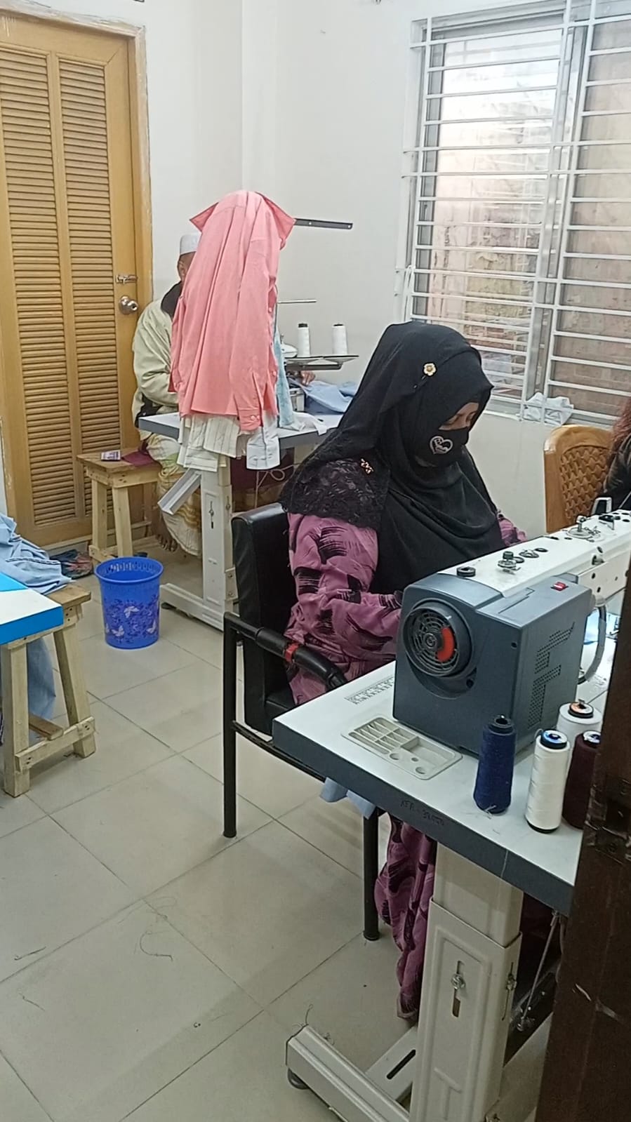 A seamstress wearing a black niqab operates an industrial sewing machine in a professional garment workshop.