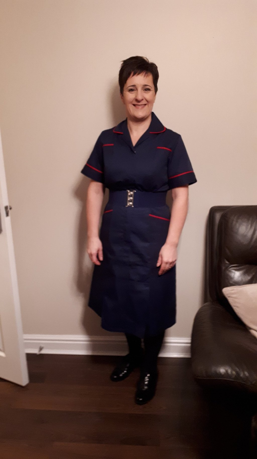 Smiling healthcare professional in a navy blue nurse uniform with red trim and a silver buckle belt.