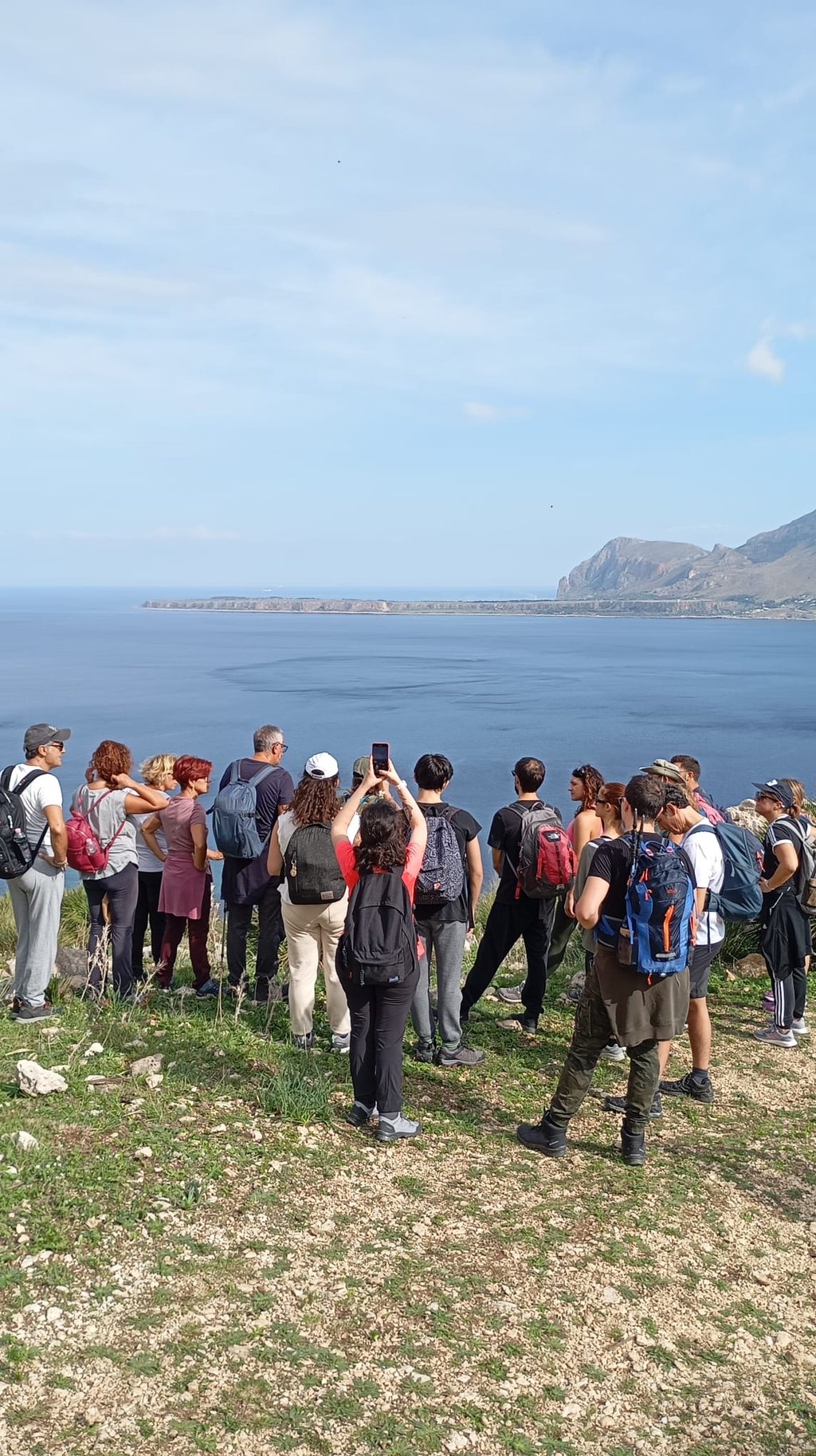 A group of hikers with backpacks overlooks the scenic blue coastline and mountains in Sicily.