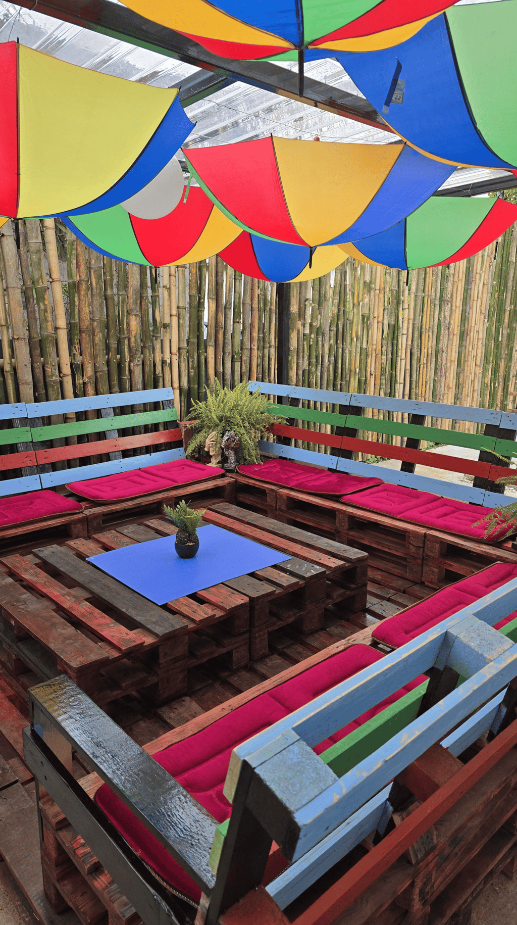 Outdoor patio lounge with colorful umbrella ceiling, wooden pallet furniture, and bamboo privacy fence.