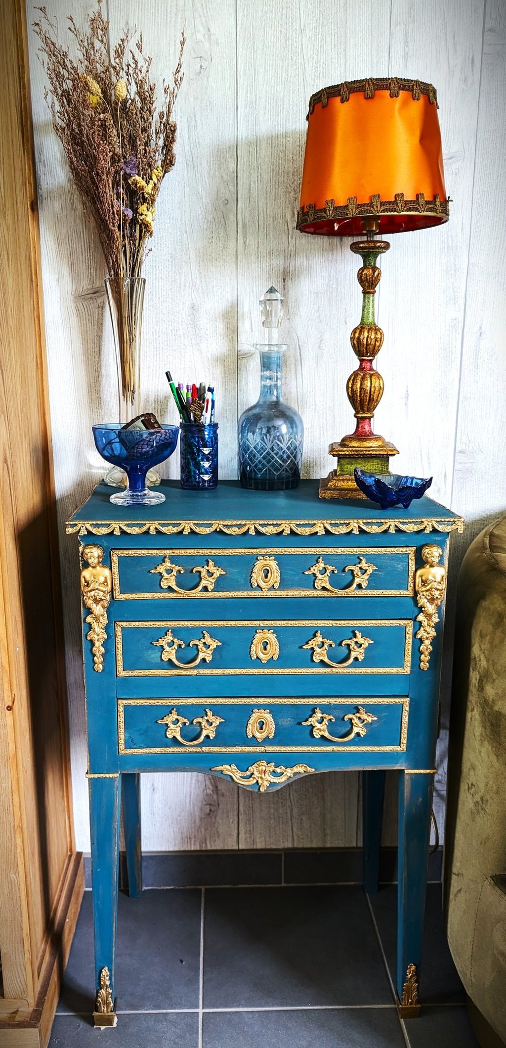 Vintage blue commode with ornate gold hardware, topped with an orange lamp and antique glassware.