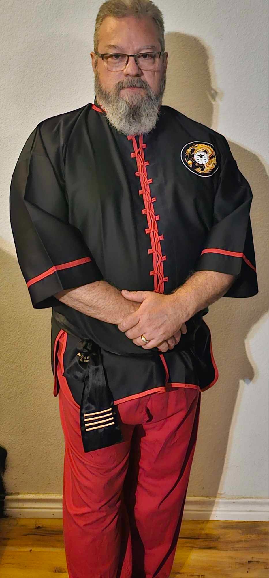 A bearded man wearing a black martial arts uniform with red trim and a gold dragon patch.
