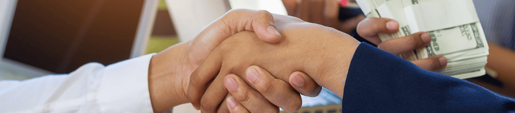 Business partners shaking hands over a contract while a woman holds stacks of US dollar bills.