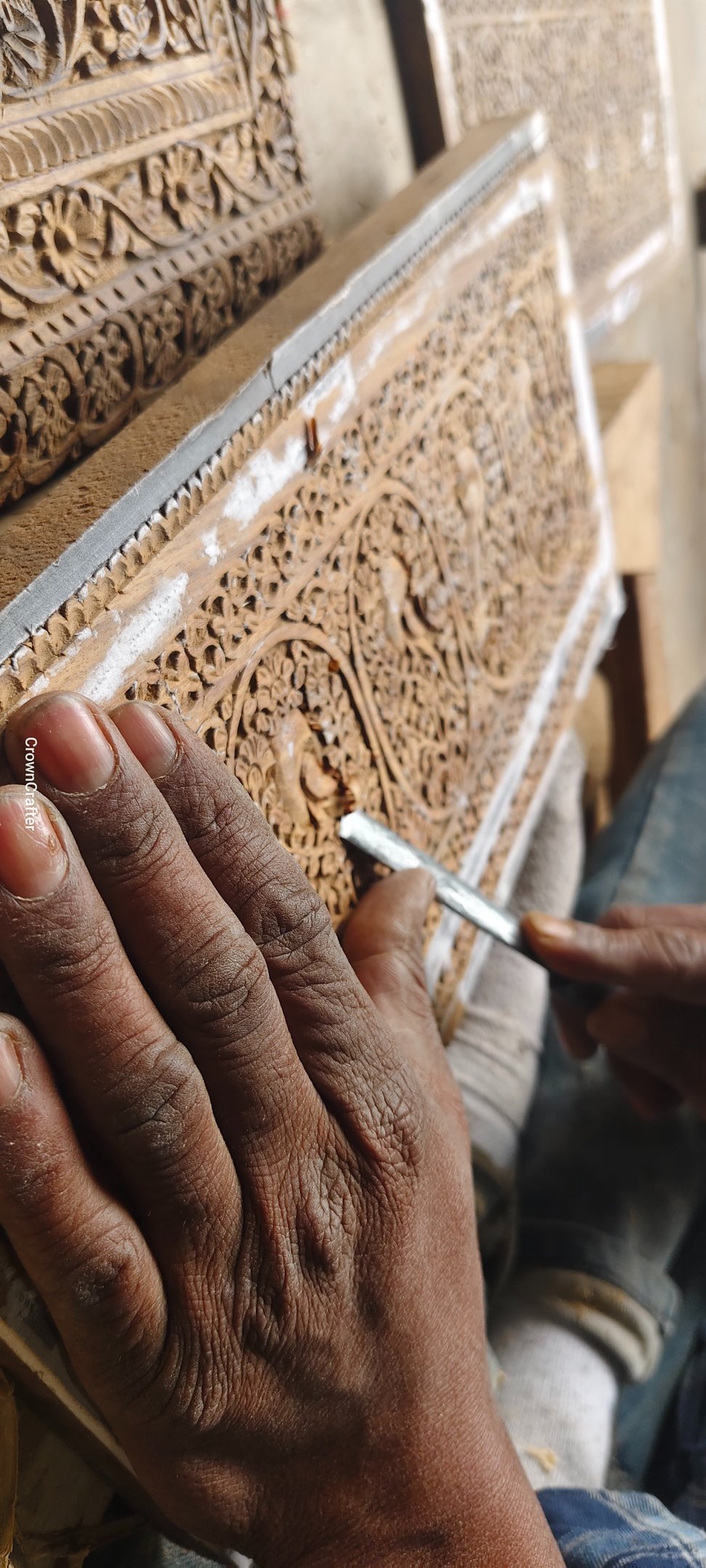 Artisan hand carving intricate floral patterns into a wooden panel using a traditional metal chisel.