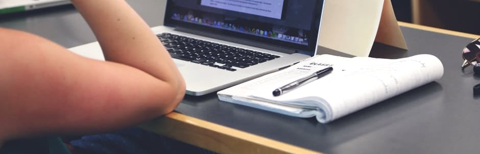Someone's elbow rests on a table next to an open notebook and laptop.