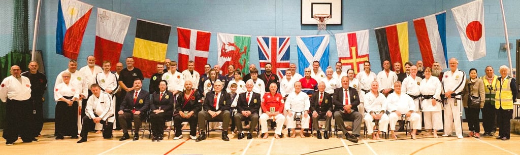 A group of martial arts students and instructors in white gi pose before international flags at a dojo.