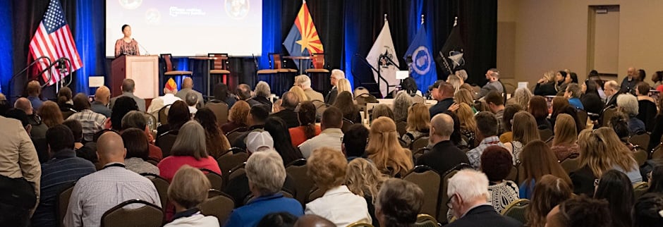 Image shows a crowded room, people in a presenation, watching a speaker ahead of them