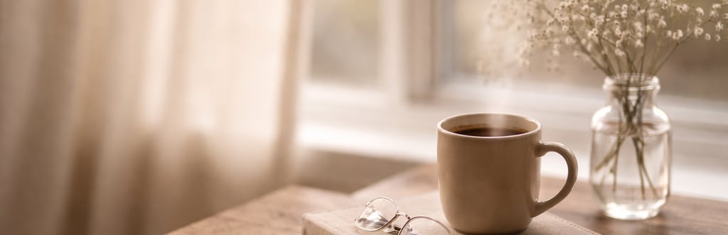 Steaming cup of coffee on a book with glasses and baby's breath flowers by a window.
