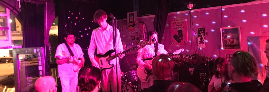 Library Siesta the band, all in white on stage playing to a packed crowd, bathed in a red light. 