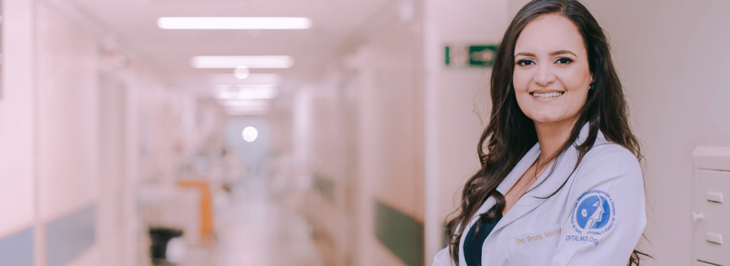 Dra Bruna Melchior de jaleco branco sorrindo de braços cruzados, aos fundos um corredor do hospital das clinicas da USP