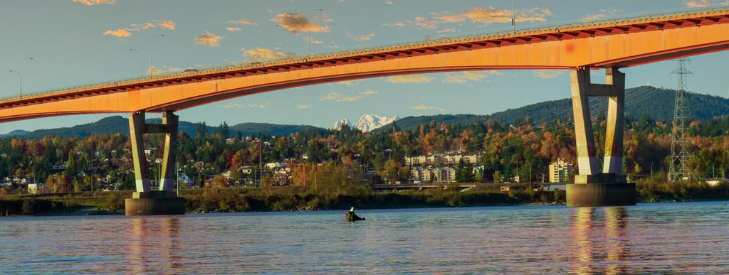 Golden Ears Bridge spanning a river, with houses and autumn-colored trees in the background, and distant snow-capped mountain