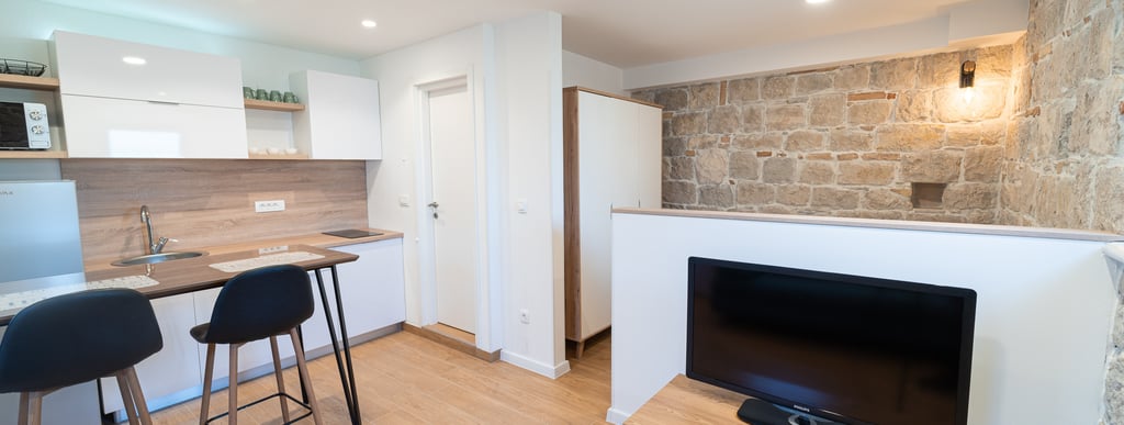 living room with walls made of stone, tv and white fully equiped kitchen