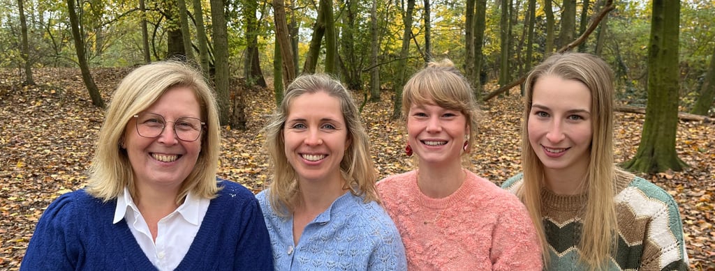 Four smiling women standing together in a scenic autumn forest with colorful fallen leaves.