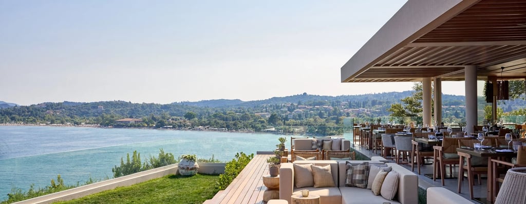 Ikos Odisia Elia restaurant terrace overlooking a turquoise sea and coastal hills.