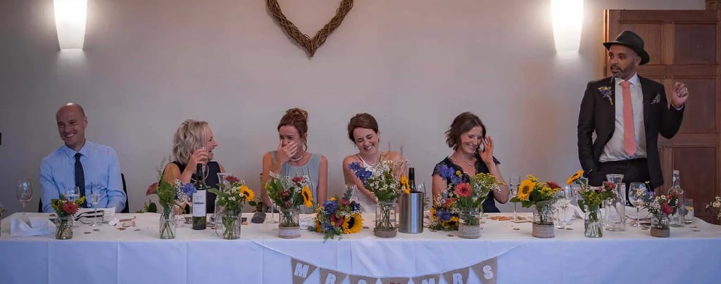 Brides and guests laughing during wedding speeches at a decorated head table with floral centerpieces.