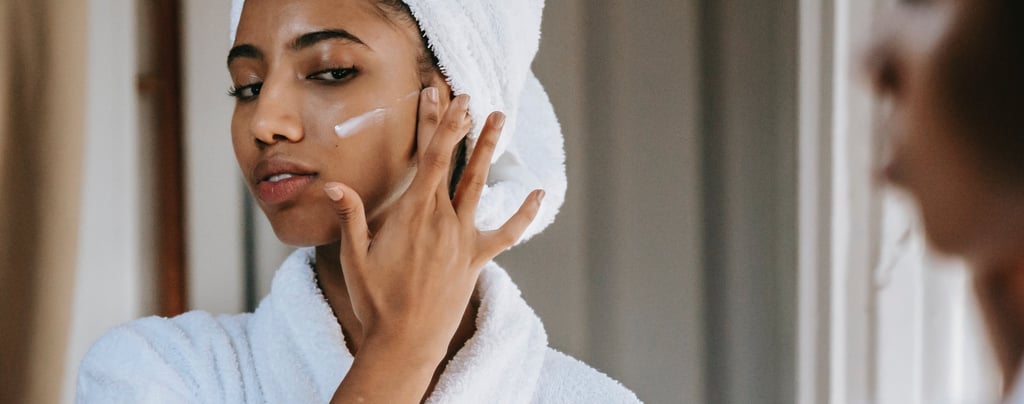 a woman in a white robe is looking at her face with a towel on her head applying face cream