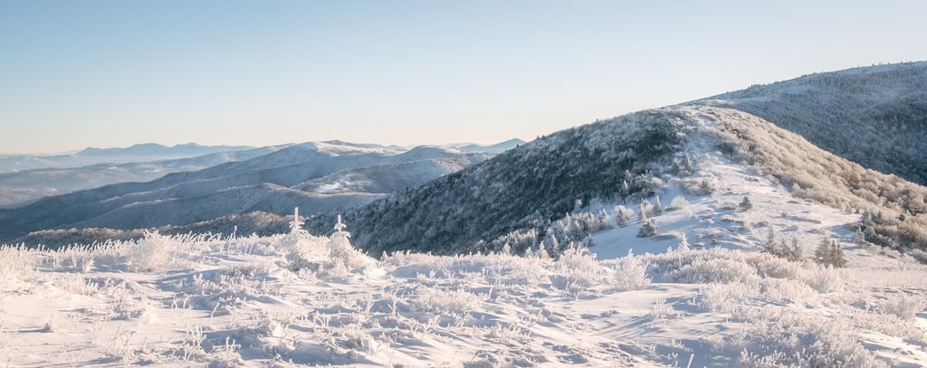 Panoramic winter landscape of snow-covered mountain peaks and frost-covered trees under a clear blue sky.