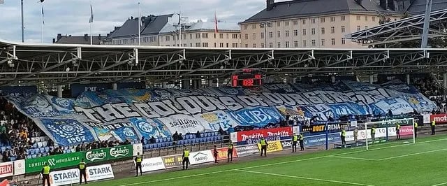 Fans von HJK Helsinki feiern in der Bolt Arena, mit blau-weißen Schals, Tribünen im Hintergrund und lebendiger Stadionstimmun