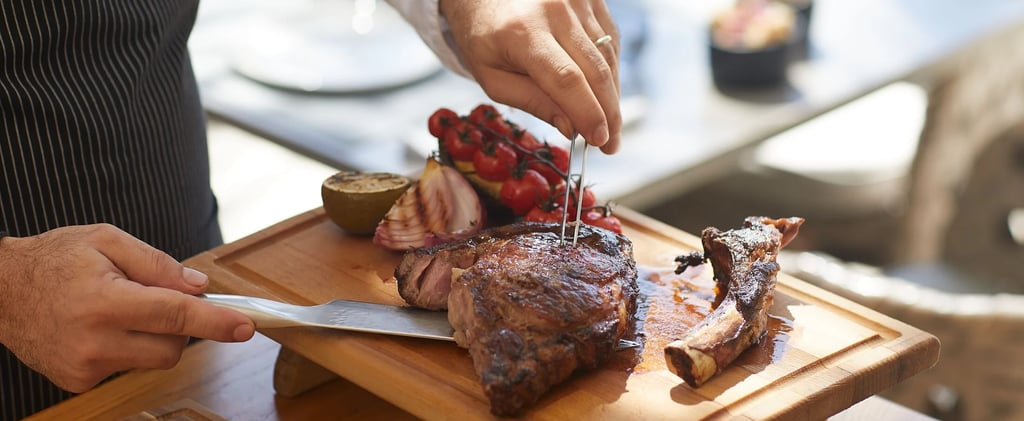 A chef carves a grilled ribeye tomahawk steak on a wooden board with roasted tomatoes and onions.