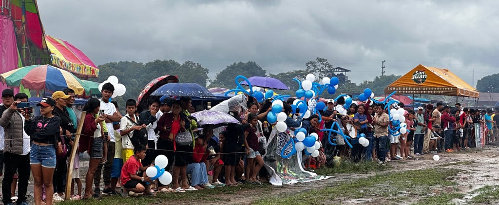 A large crowd gathers on a muddy field under a cloudy sky for an outdoor community festival celebration.