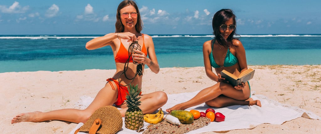 Two young women in bikinis enjoy a tropical beach picnic with fresh fruit and books.